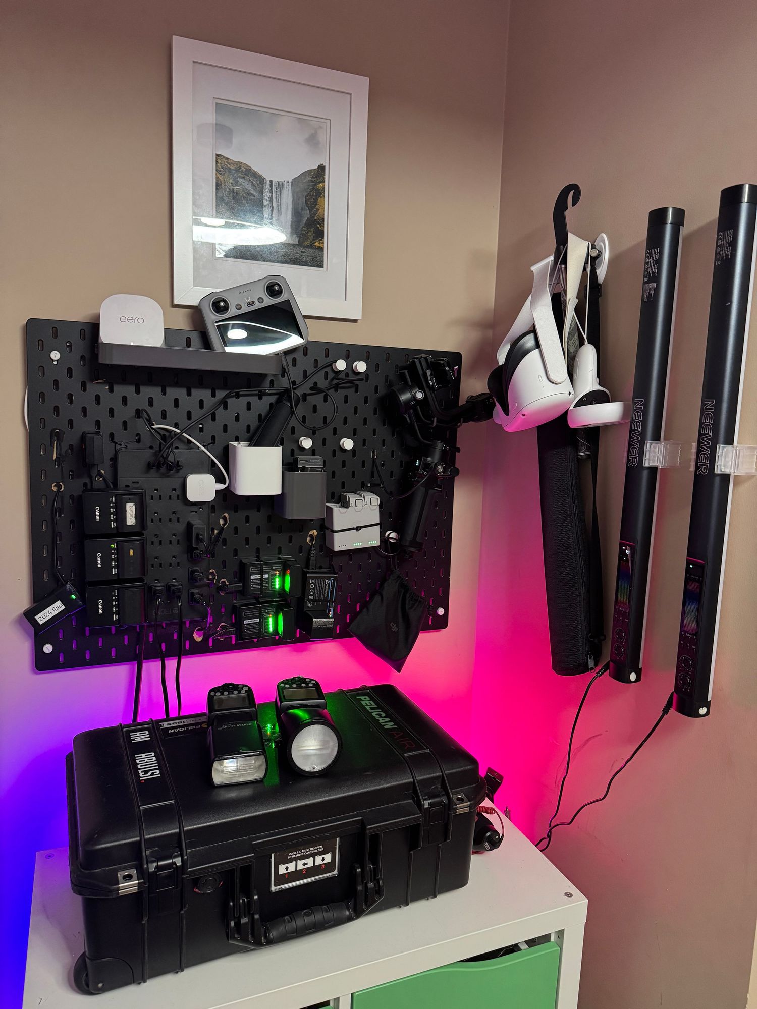 A pegboard organizer with charging cables and electronics above a Pelican case, with colorful RGB lighting in a home setup.