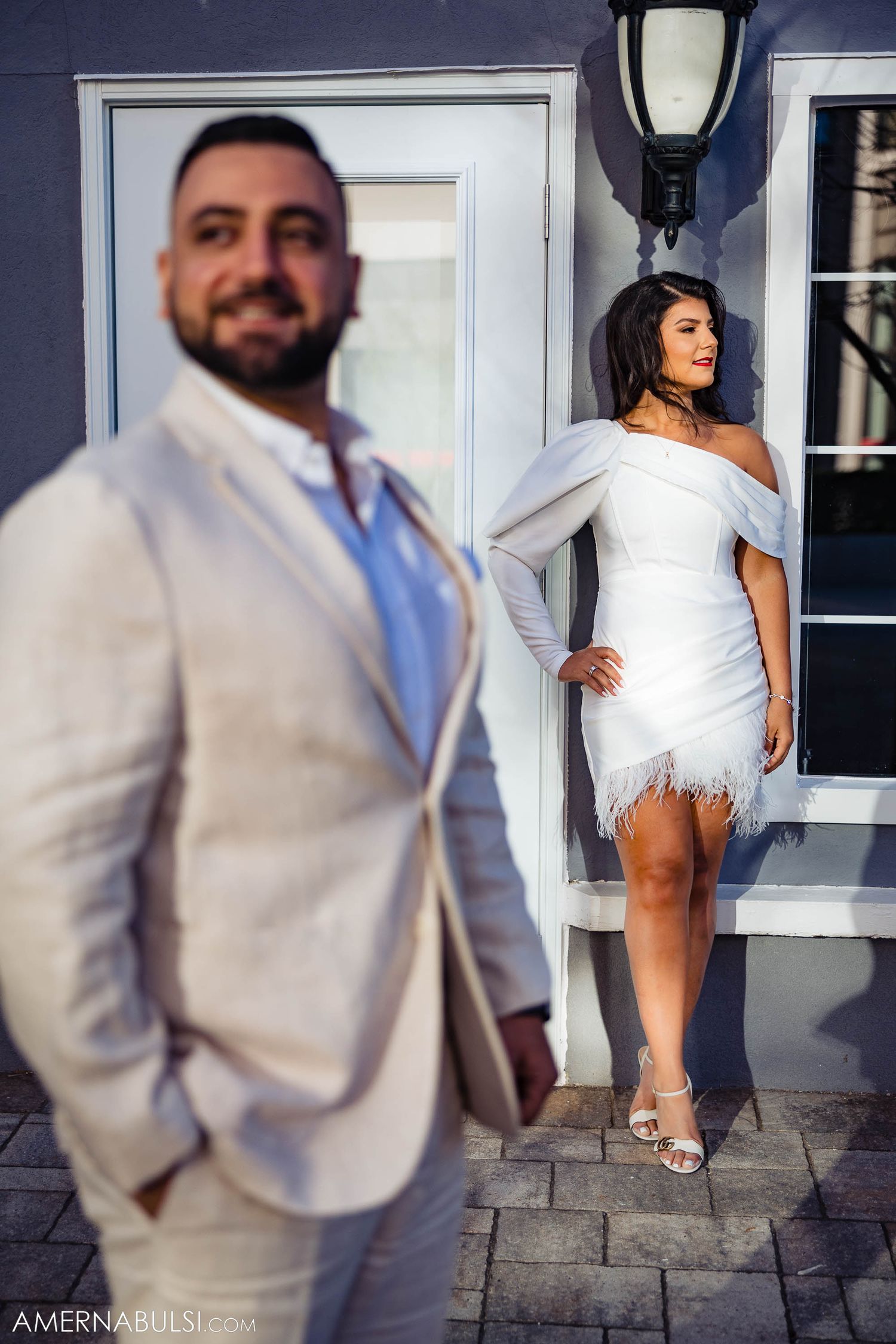 A stylish couple in white and beige formal attire posing separately against a blue exterior wall.