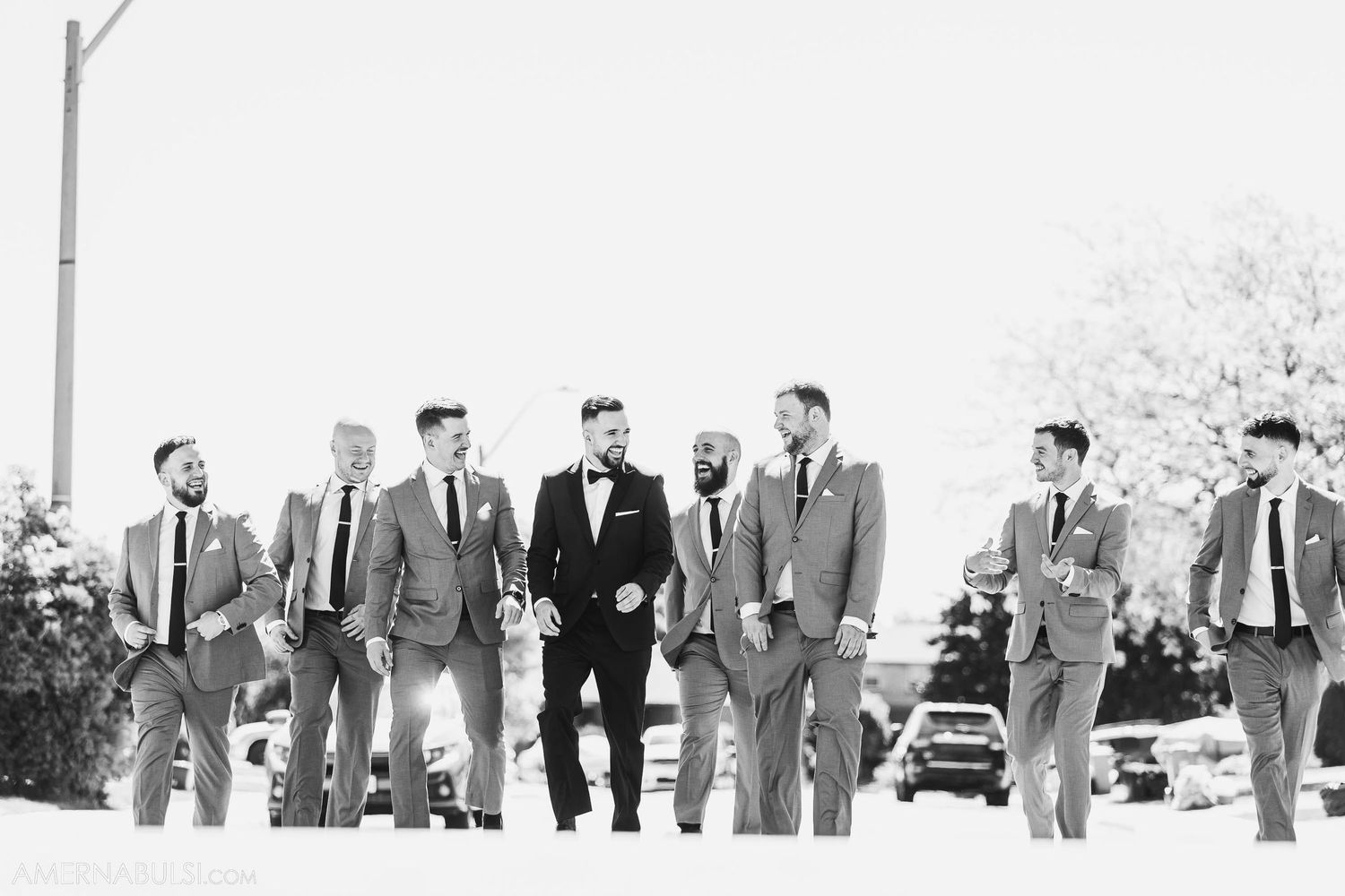 A wedding party walks together outdoors with the groom in black tuxedo centered among groomsmen in gray suits.