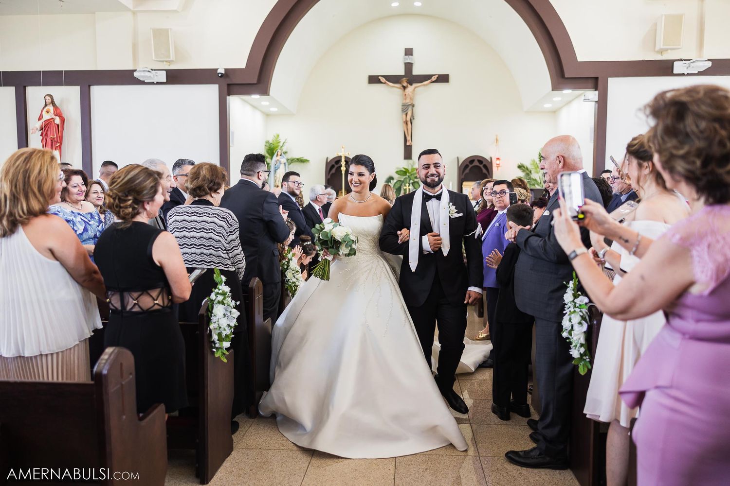 Wedding recessional with guests taking photos as couple walks down church aisle.