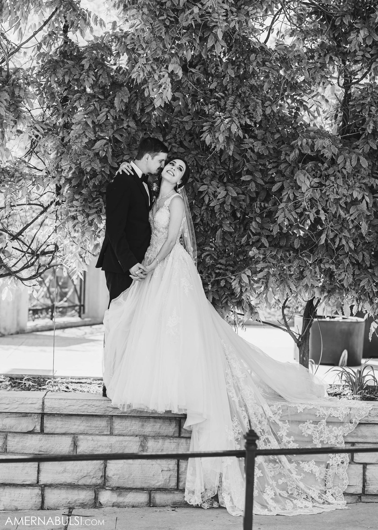 A romantic black and white wedding photo of a couple posed against lush foliage on stone steps.
