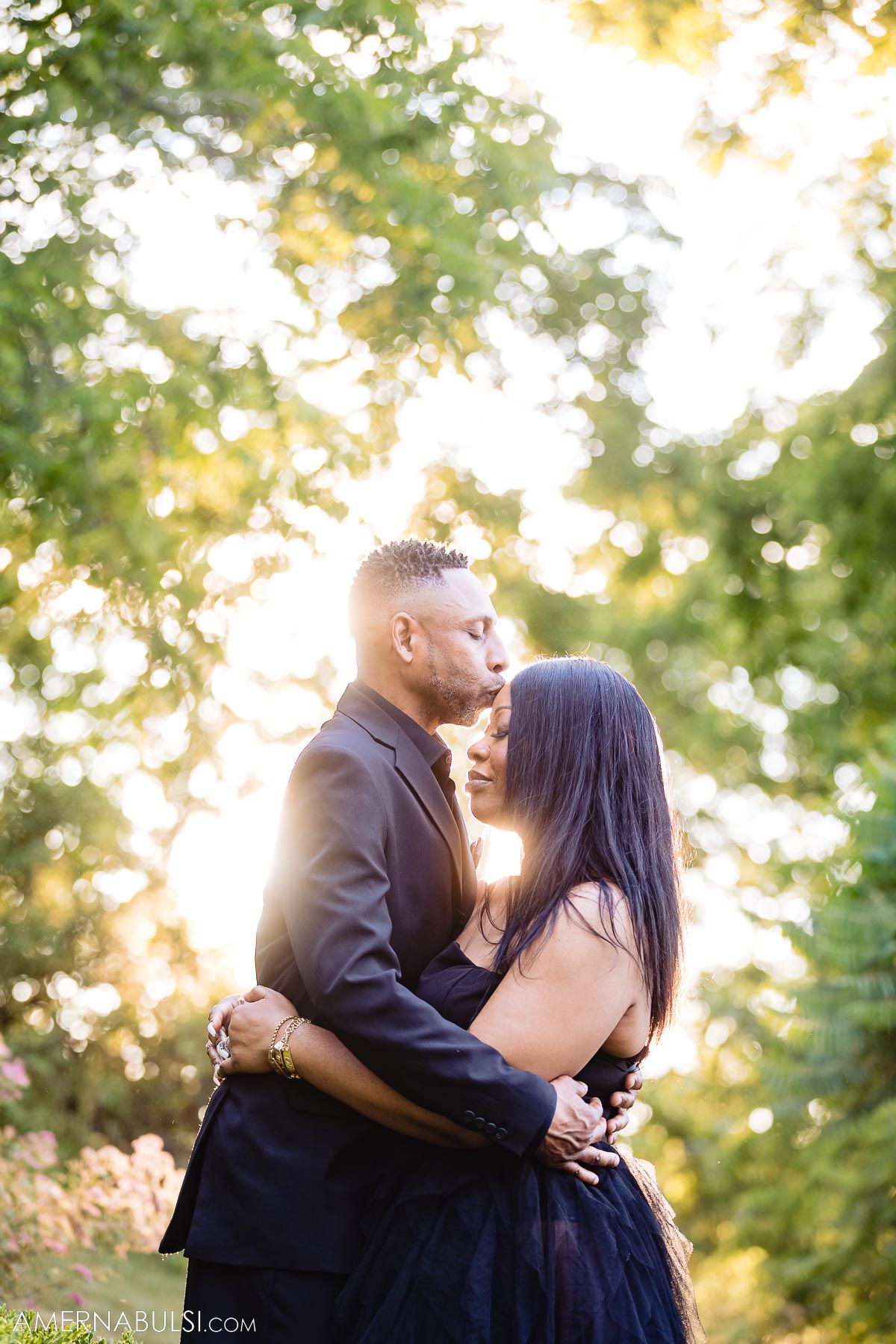 Couple embraces during sunset with golden light filtering through trees in the background.
