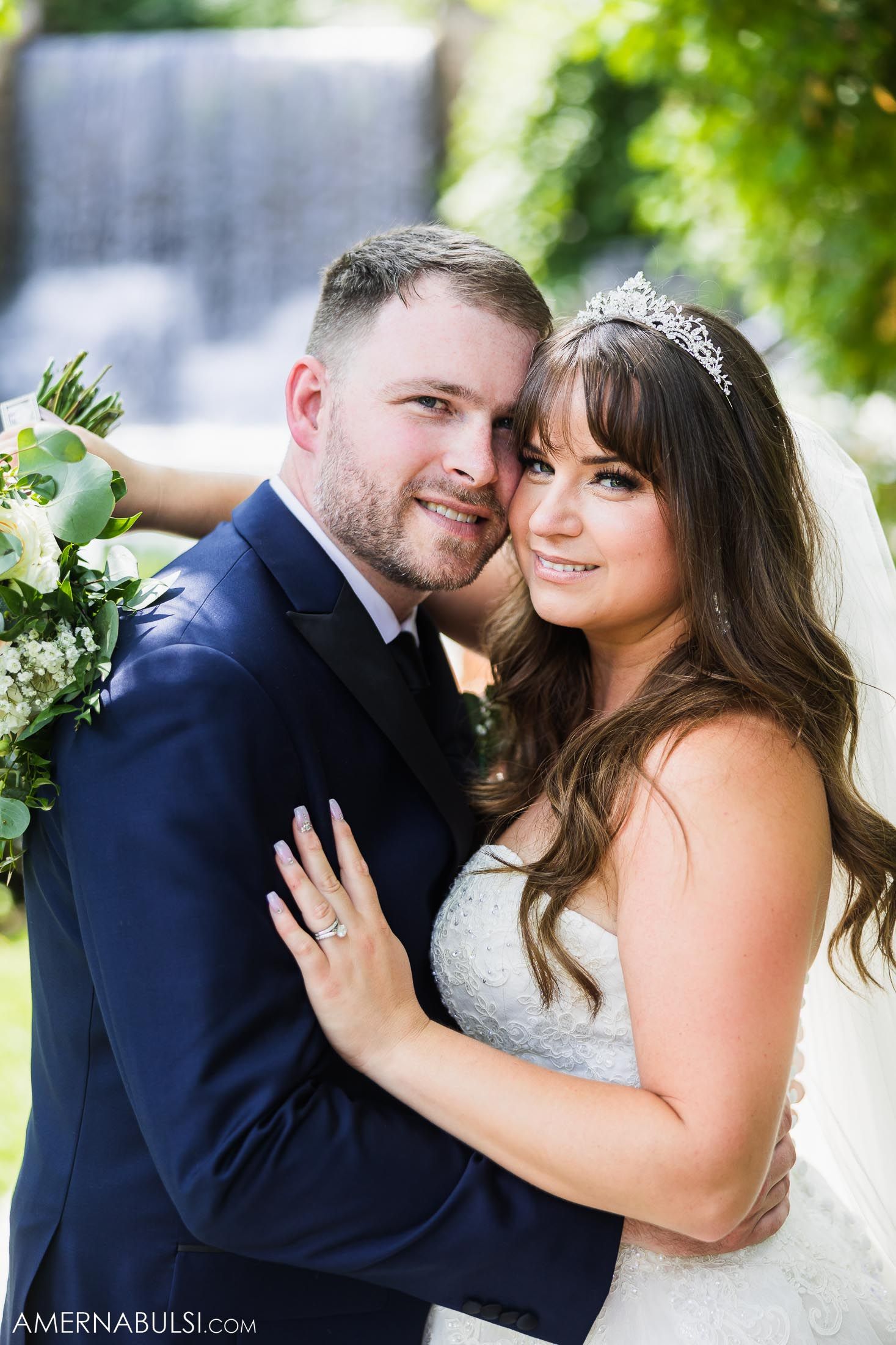 Elegant wedding portrait of couple in navy suit and white gown with waterfall backdrop.
