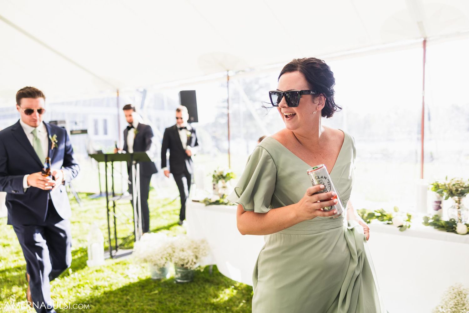 Wedding guests in formal attire mingle under a white tent during an outdoor celebration.