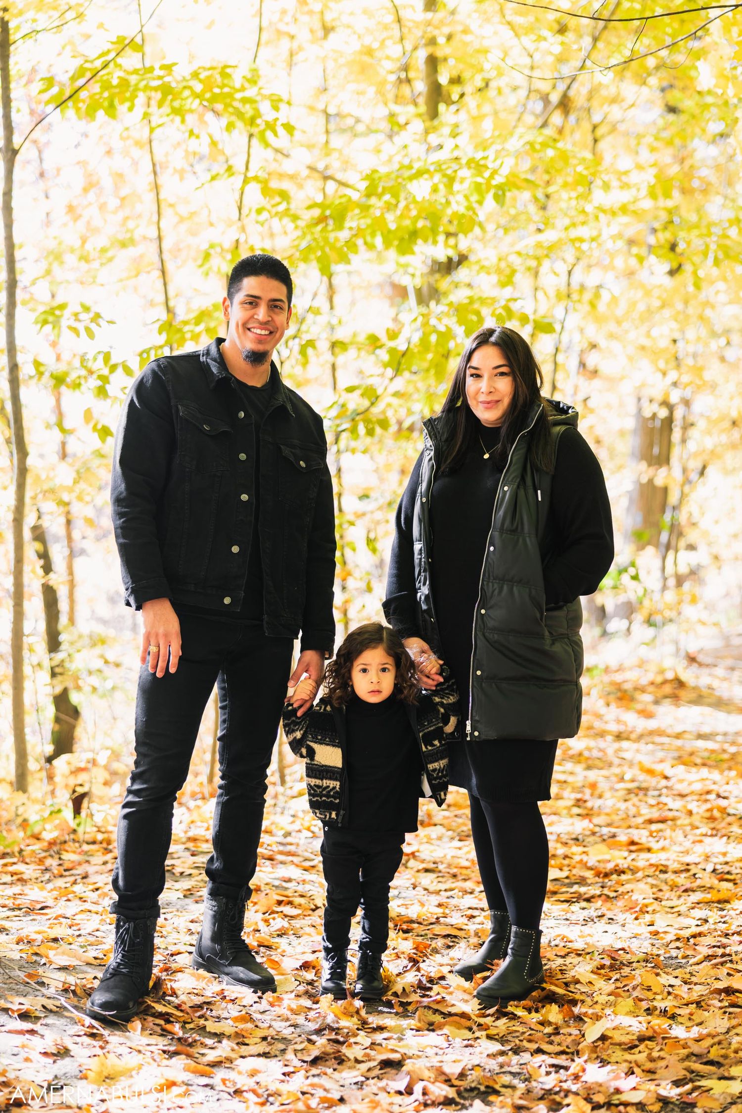 A family poses together in matching dark outfits against a backdrop of yellow autumn trees.