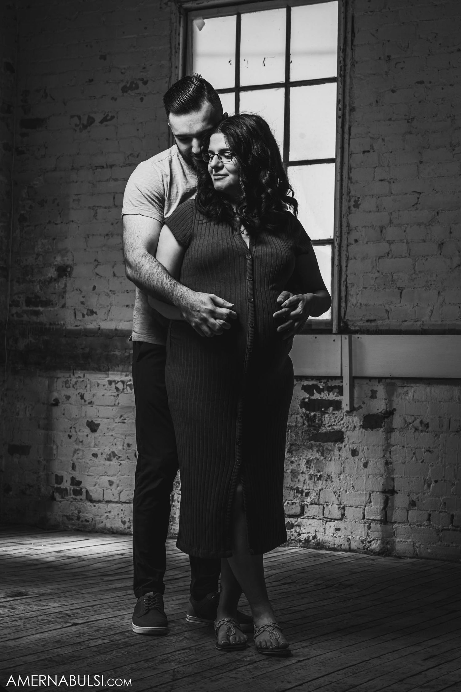Black and white maternity portrait against rustic brick wall and window.