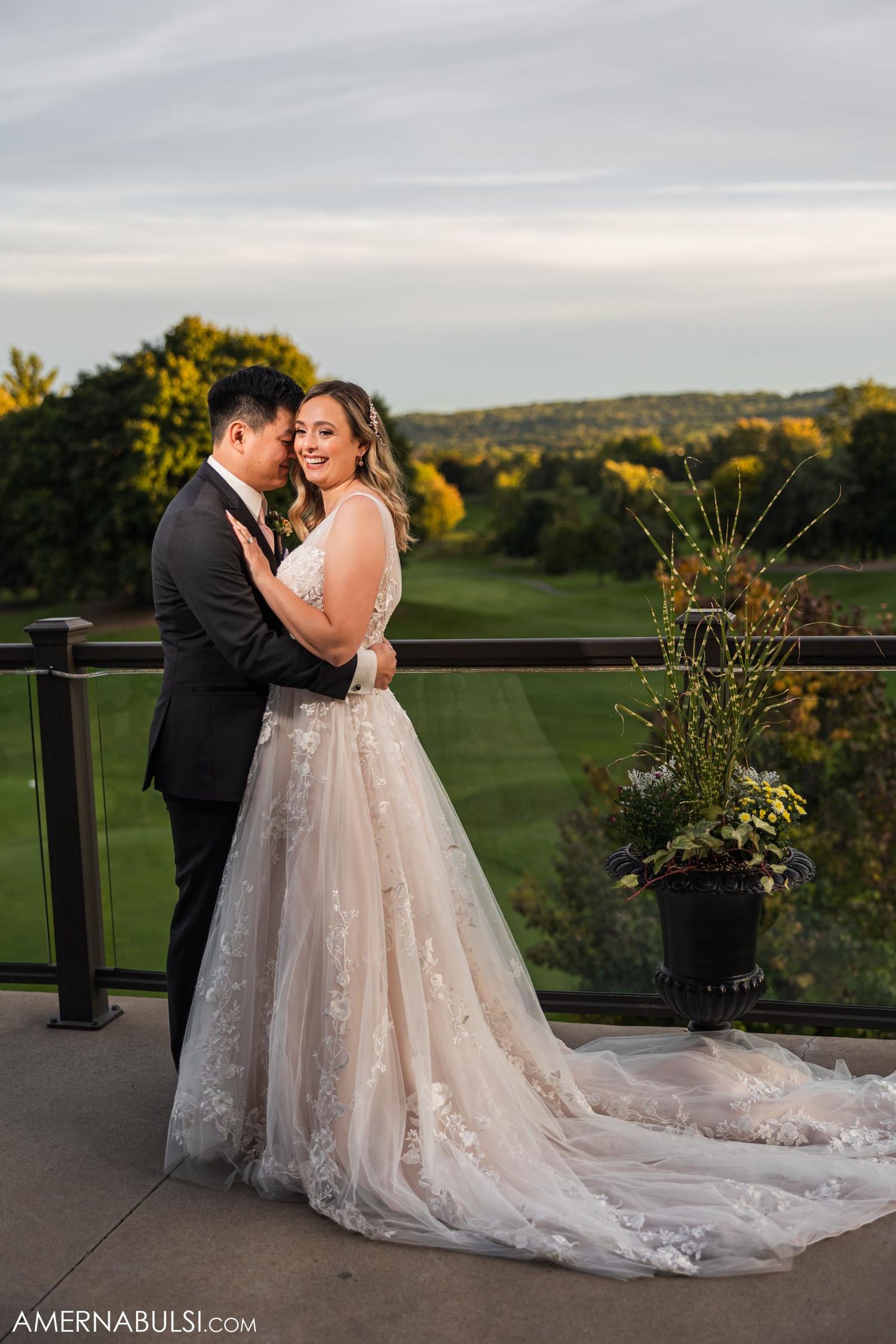 Newlyweds embrace on outdoor terrace with scenic rolling hills in background at sunset.