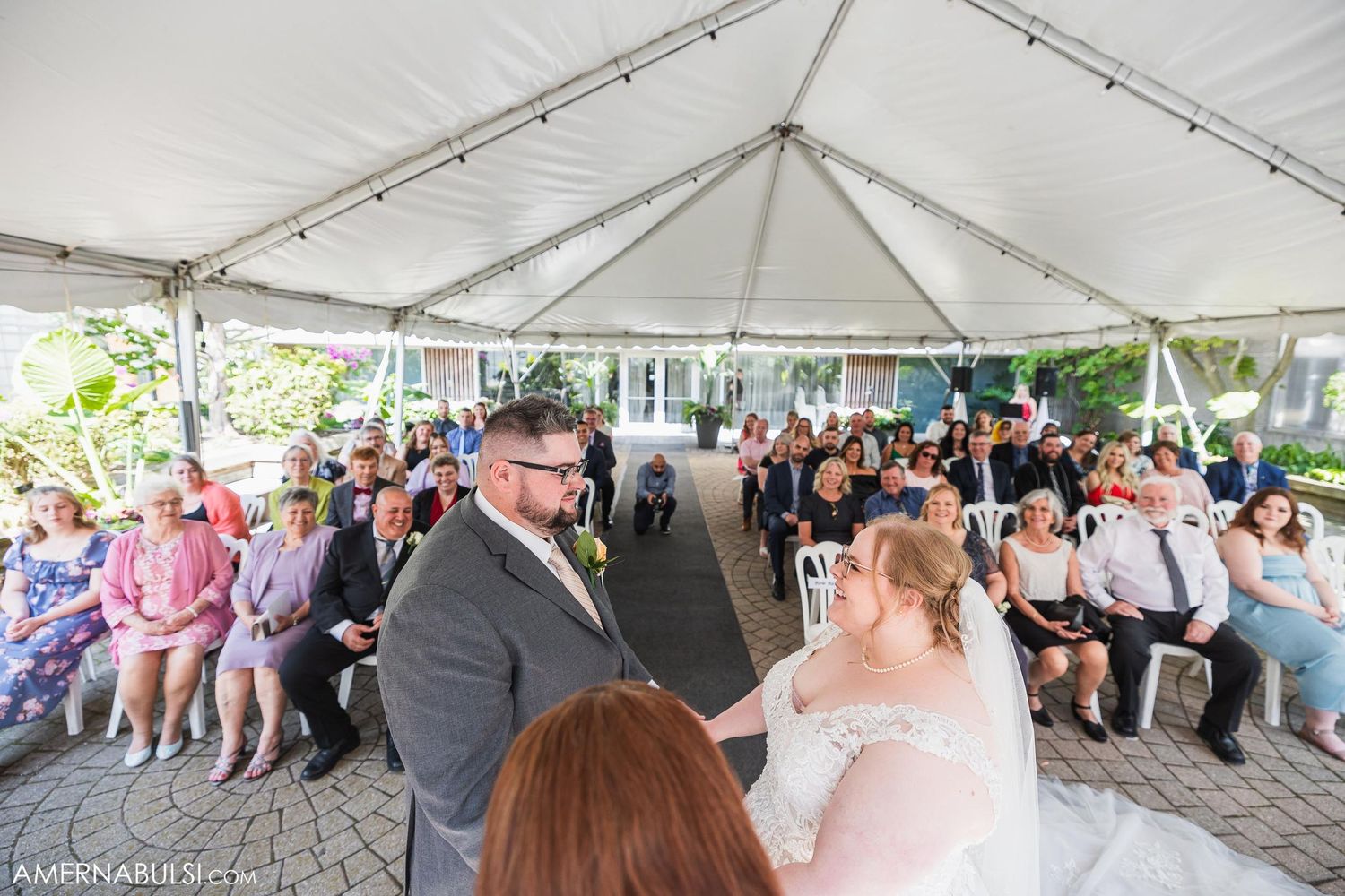 Wide view of tent wedding ceremony with guests seated on white chairs.