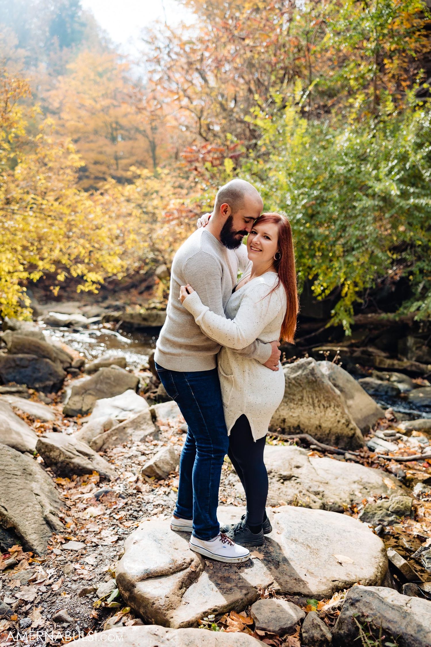 Couple embraces while standing on rocks near a stream surrounded by autumn foliage.
