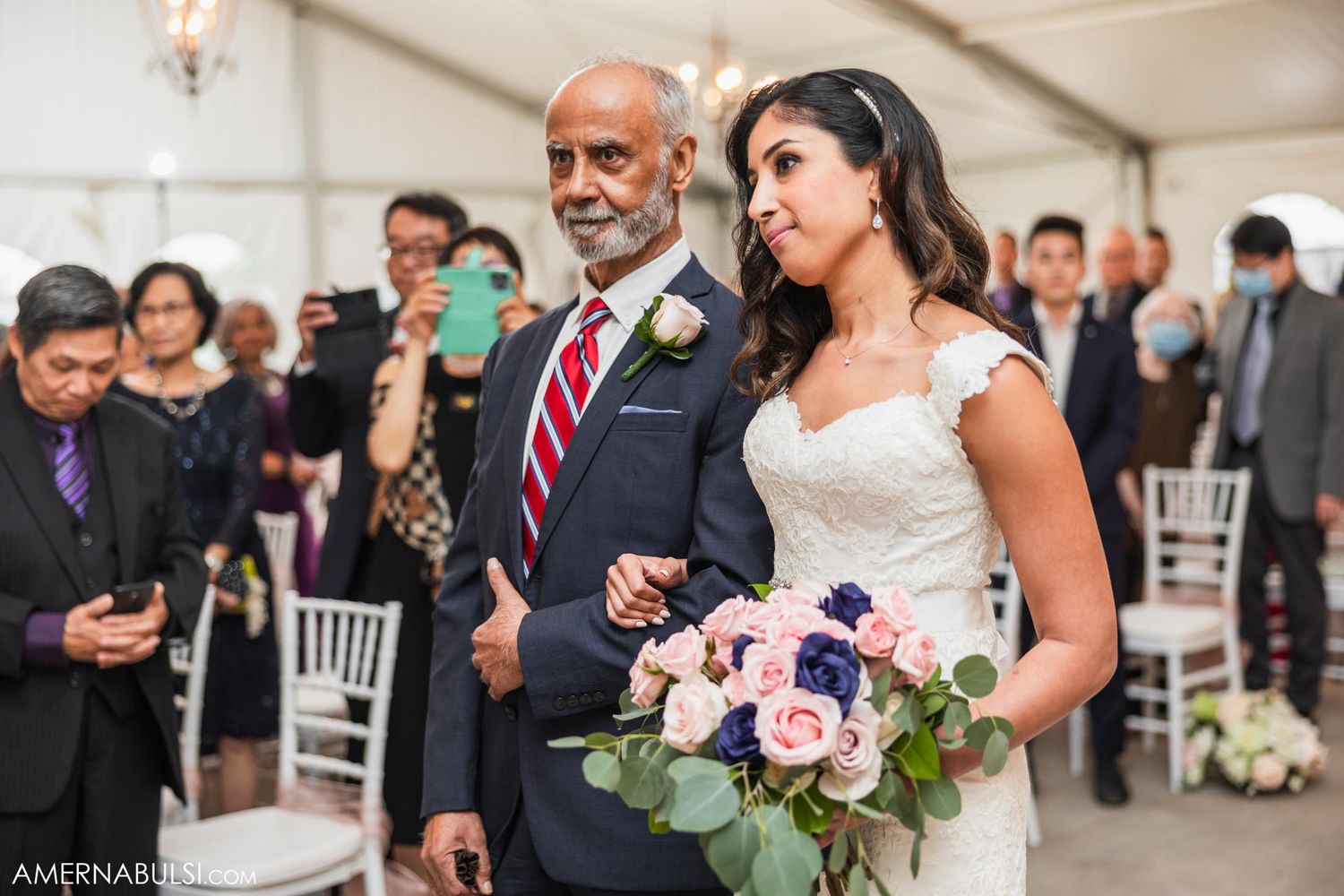 A traditional wedding ceremony takes place in an elegant white tent with pink and blue floral bouquets.