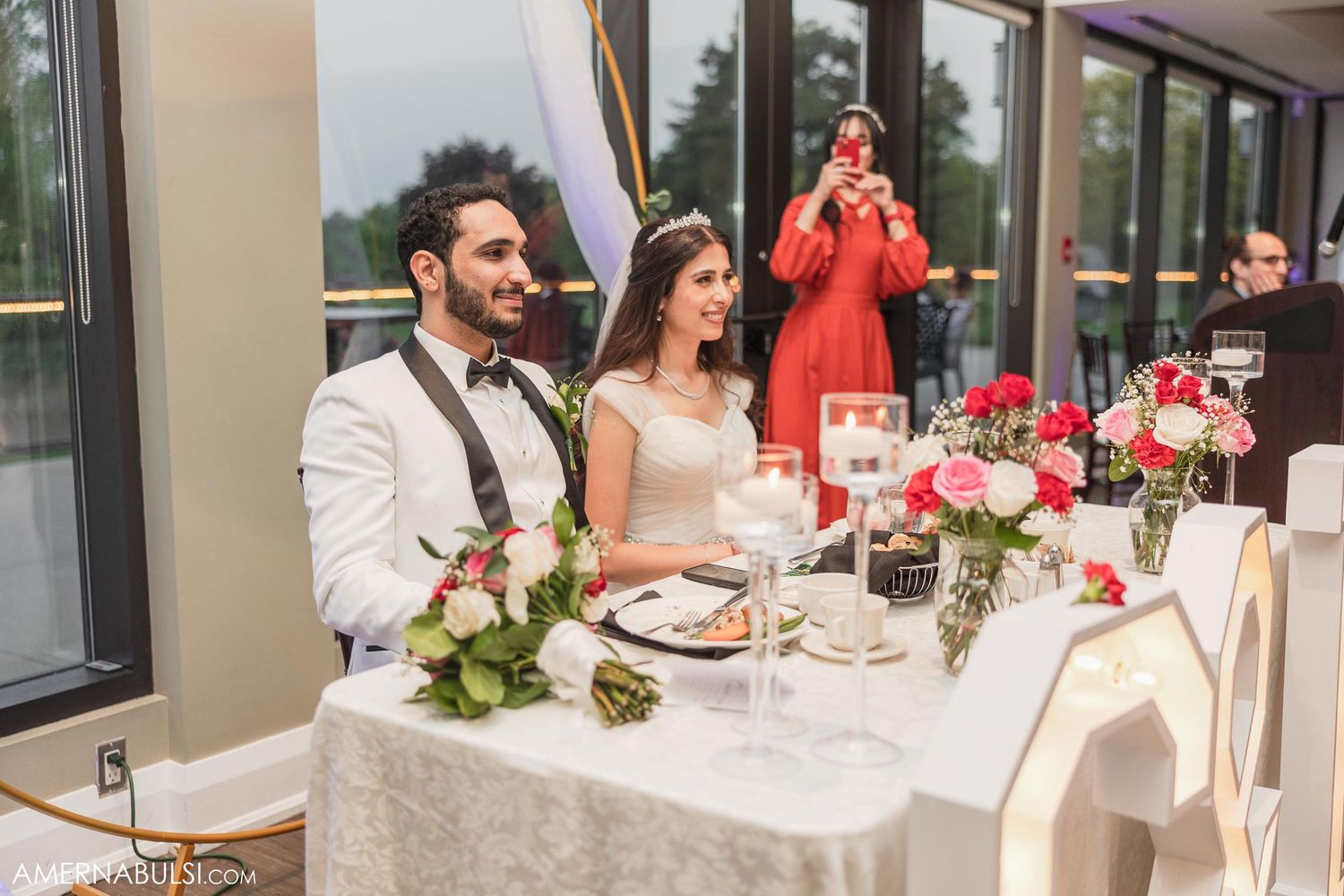 Newlyweds sit at their reception table decorated with floral centerpieces and elegant table settings.
