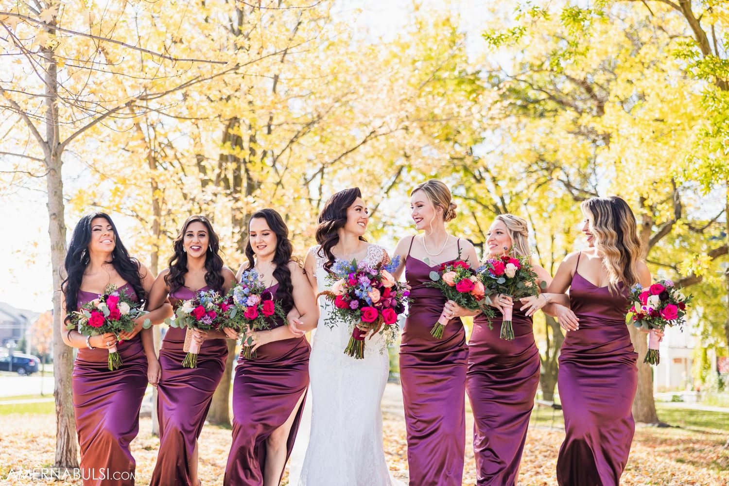 A bridal party in burgundy dresses laughs together outdoors during autumn.