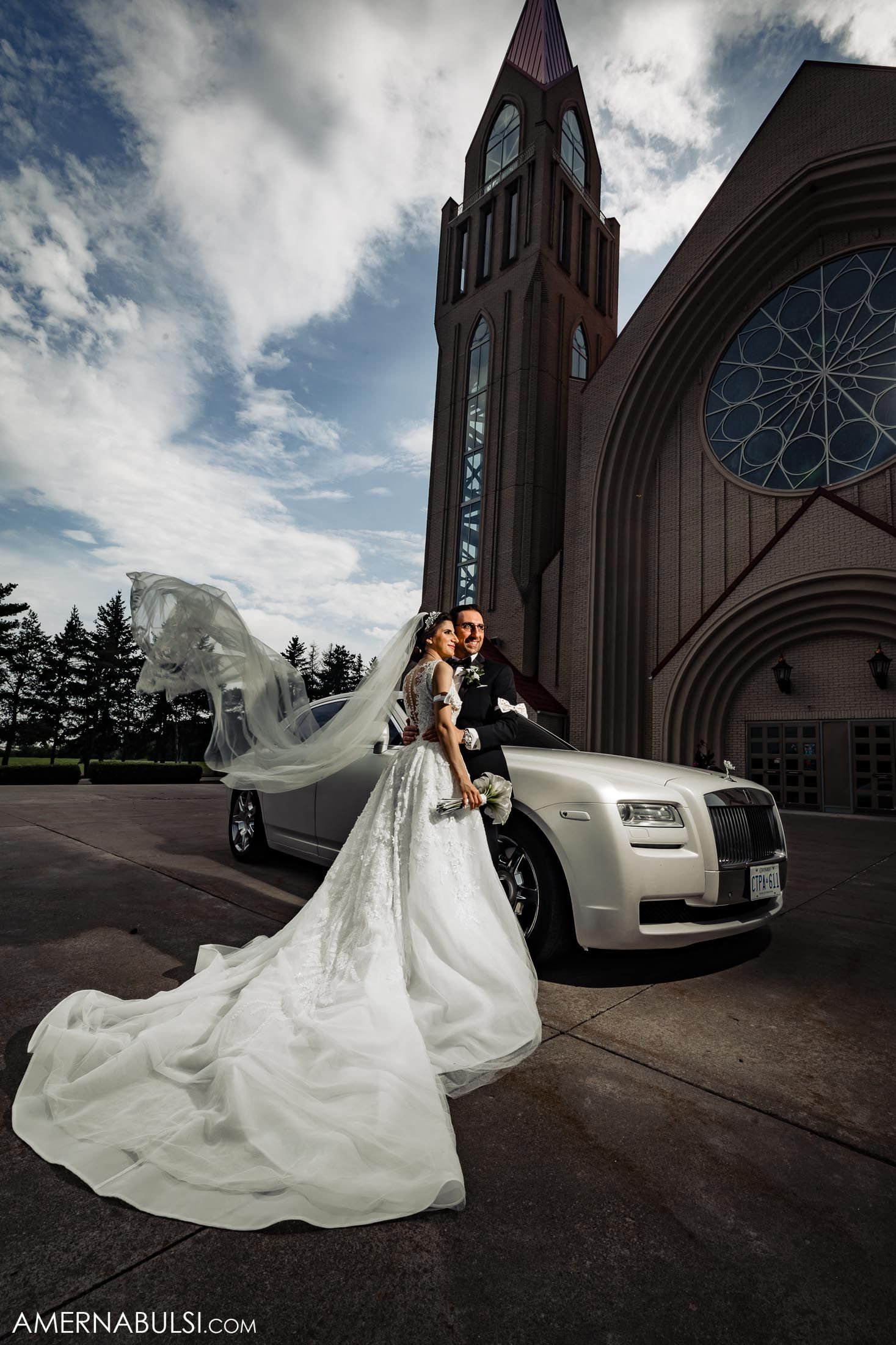 Dramatic wedding photo with flowing veil and white Rolls Royce in front of church architecture.