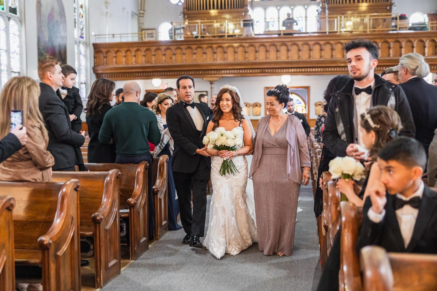 Wedding party processes through ornate church interior with wooden pews and decorative architecture.