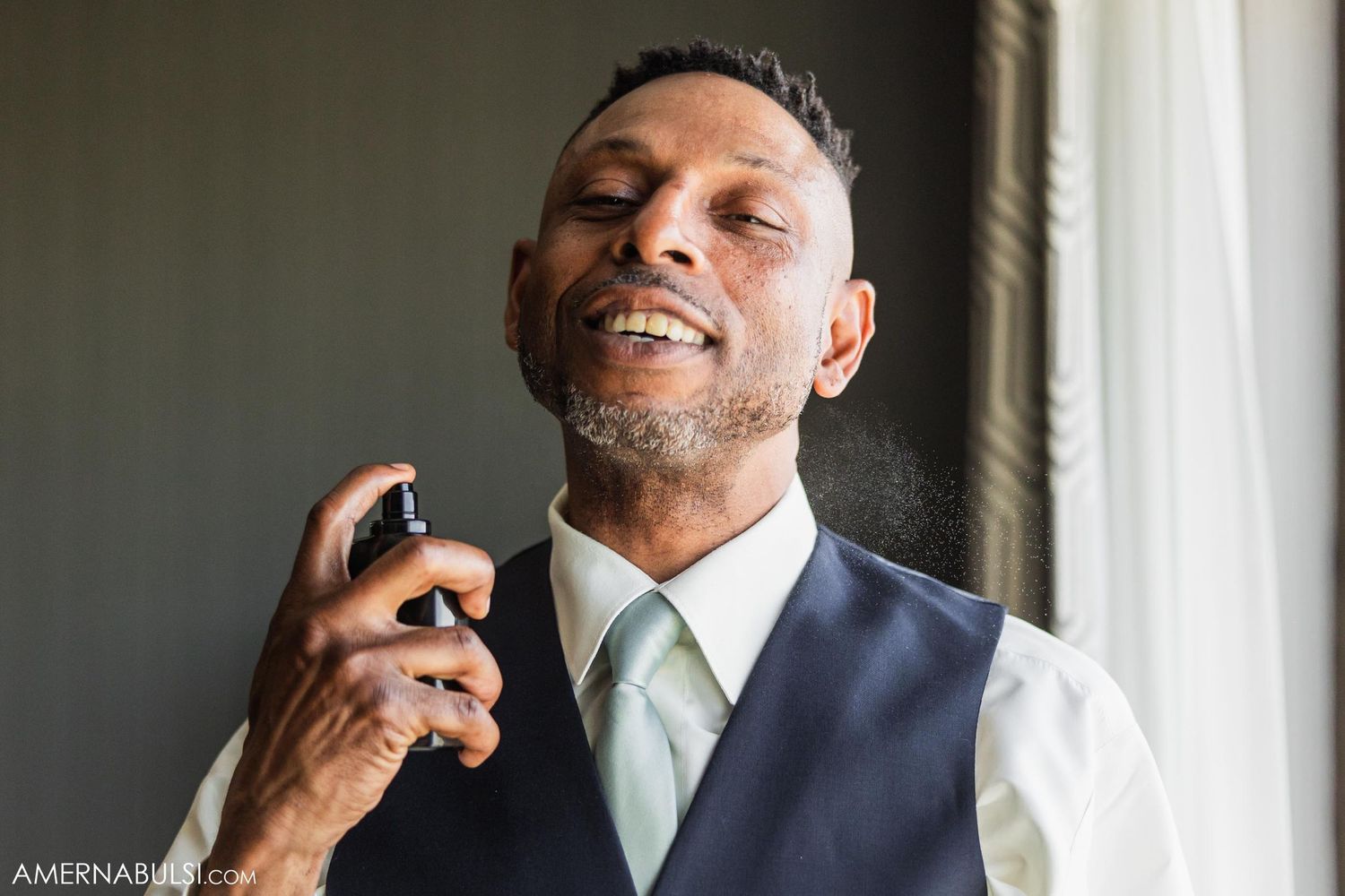 A groom in a navy vest and tie applies cologne during wedding preparations.