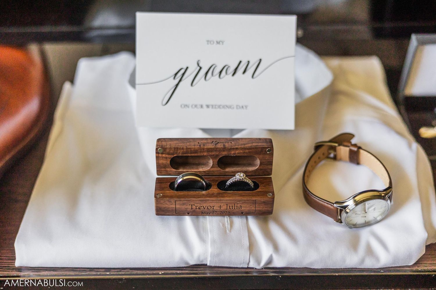 Wedding accessories including groom card, wooden ring box, and watch on white surface.