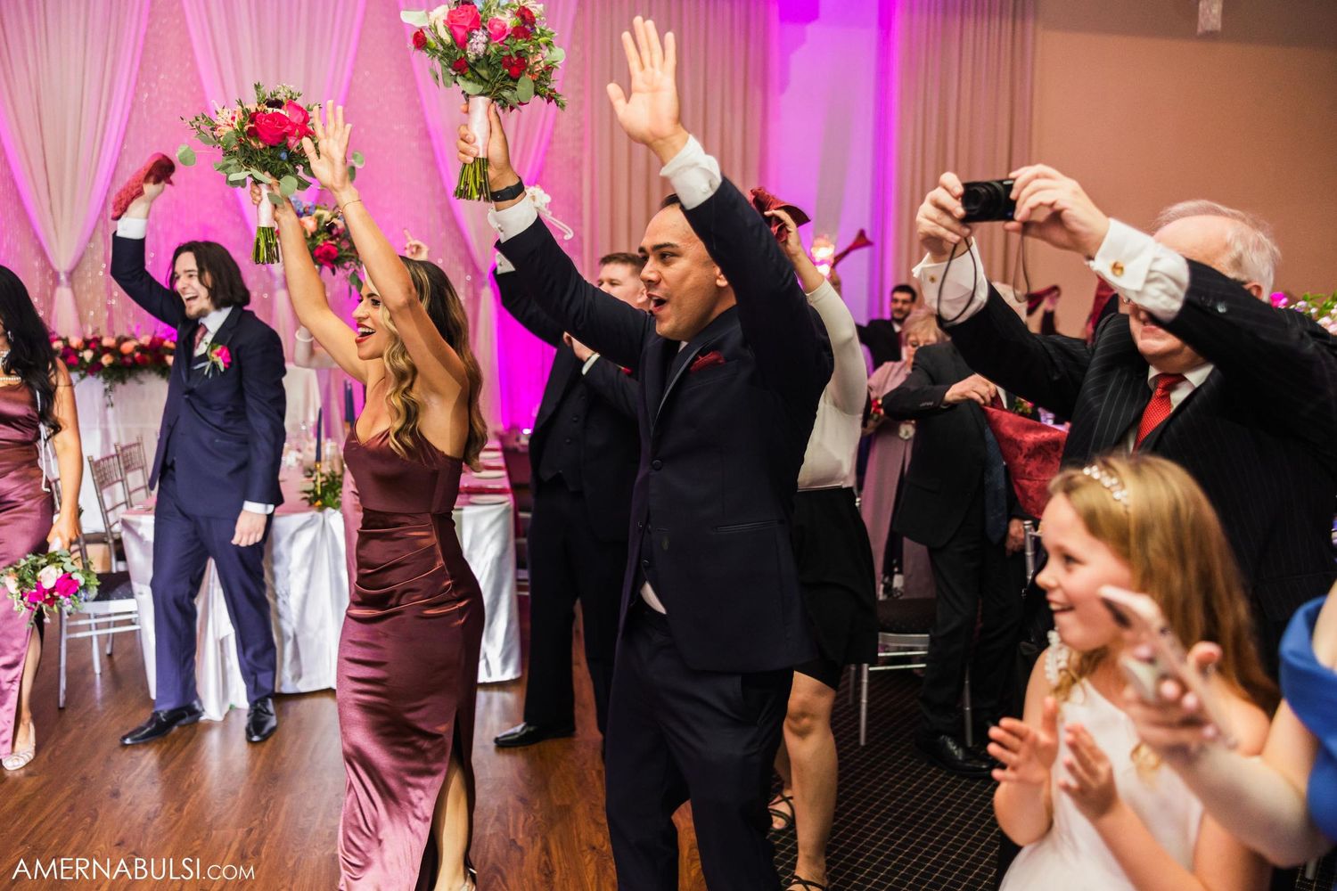 Wedding party and guests raising hands in celebration during reception with pink accented decor.