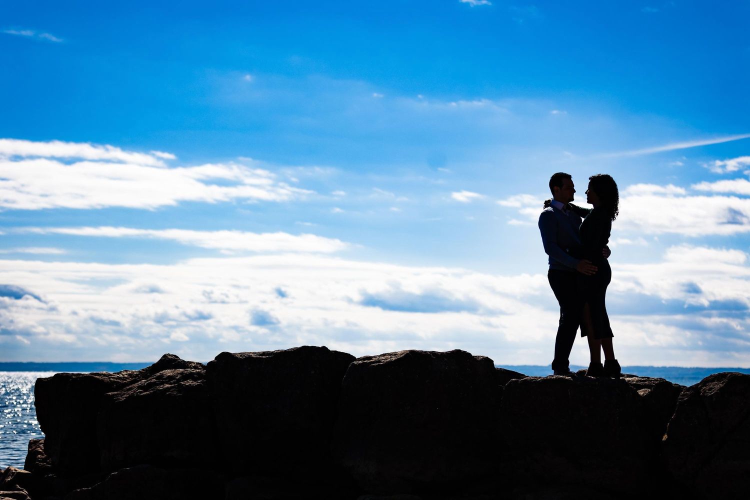 A silhouetted couple stands on rocky cliff overlooking ocean at sunset.