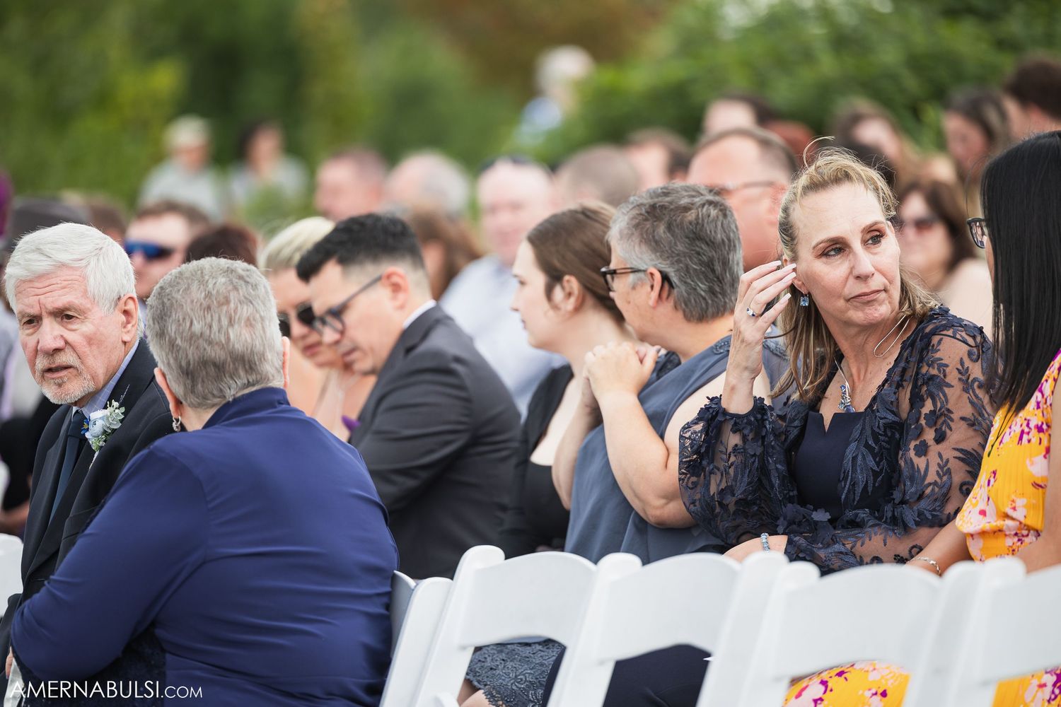 Wedding guests gather outdoors for the ceremony under green trees and natural lighting.