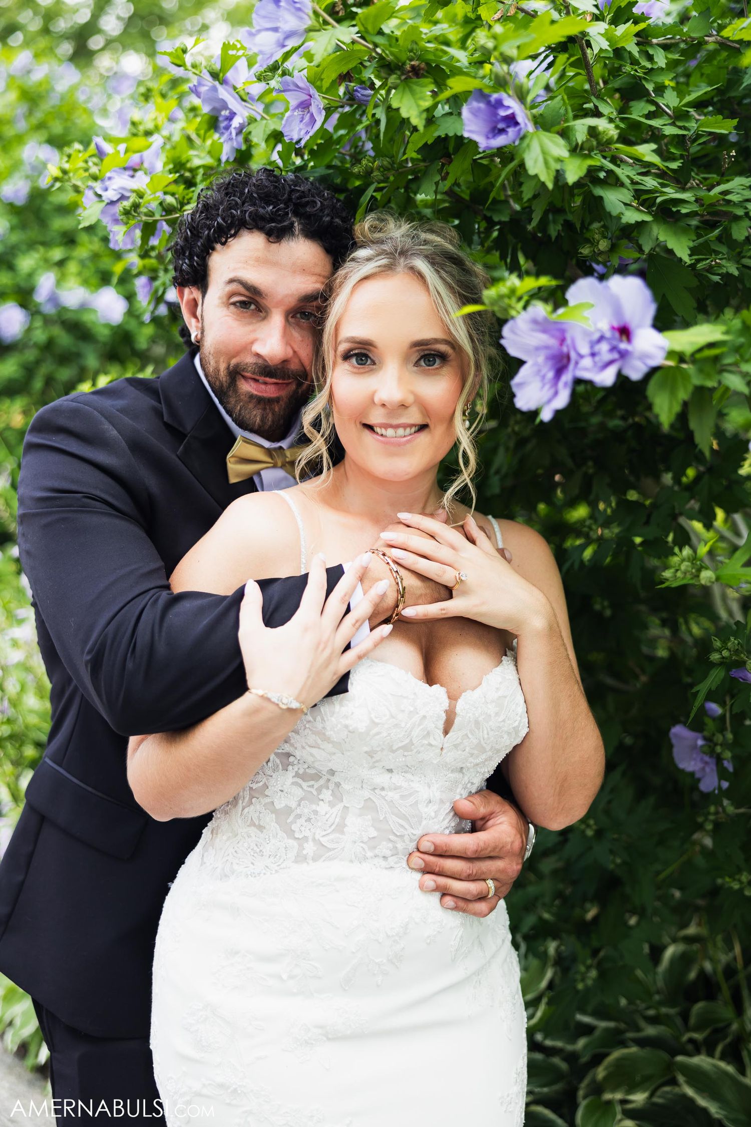 A romantic wedding portrait in a garden with purple flowers showing a tender embrace and display of rings.