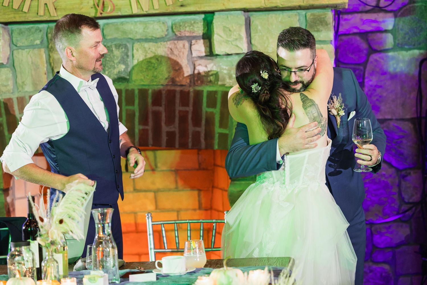 A couple embraces during their wedding reception in front of a rustic stone wall with colorful mood lighting.