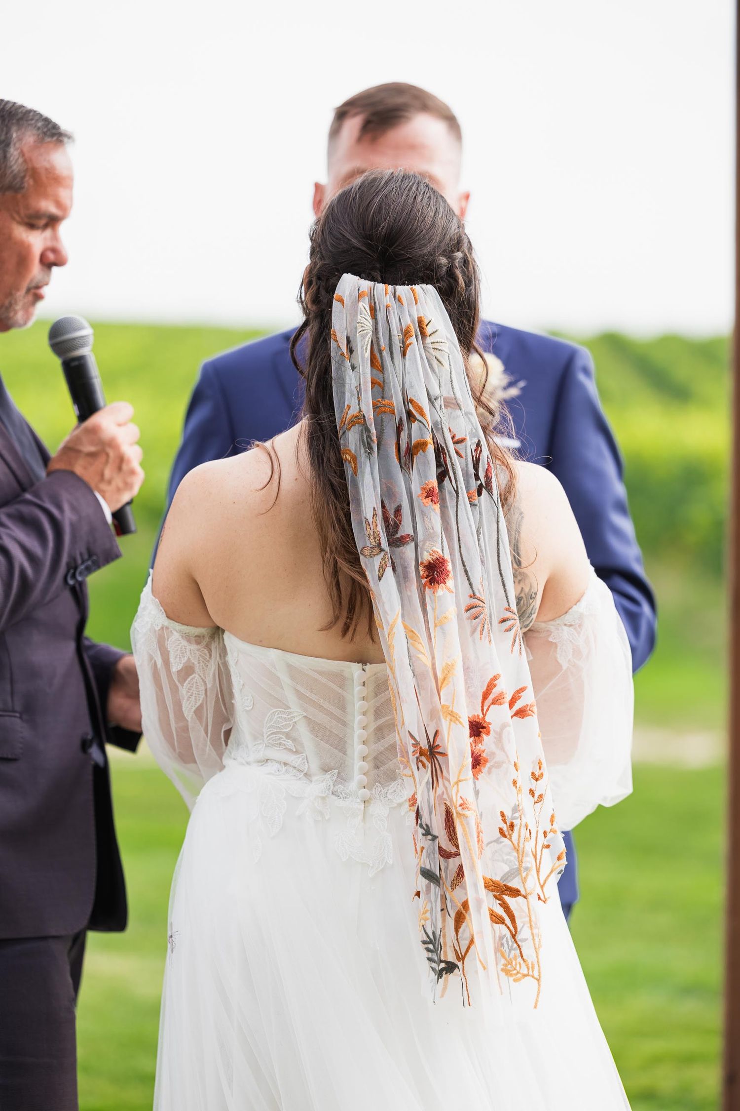 The back of a bride's dress and floral veil detail during an outdoor wedding ceremony.