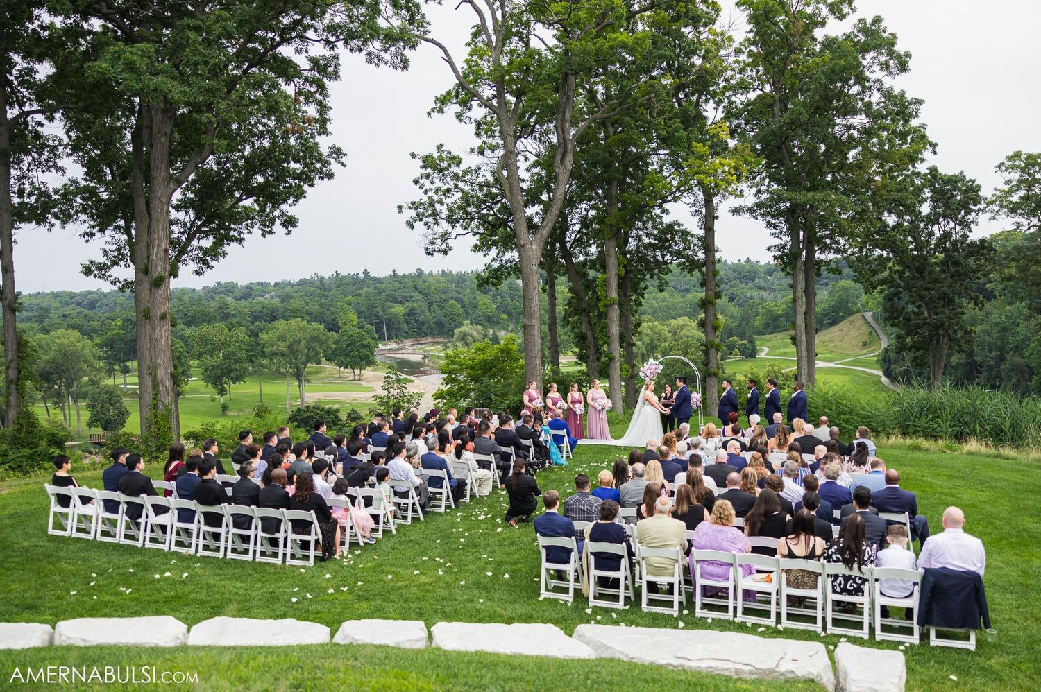 Outdoor wedding ceremony with white chairs arranged on a lush green hillside surrounded by tall trees.