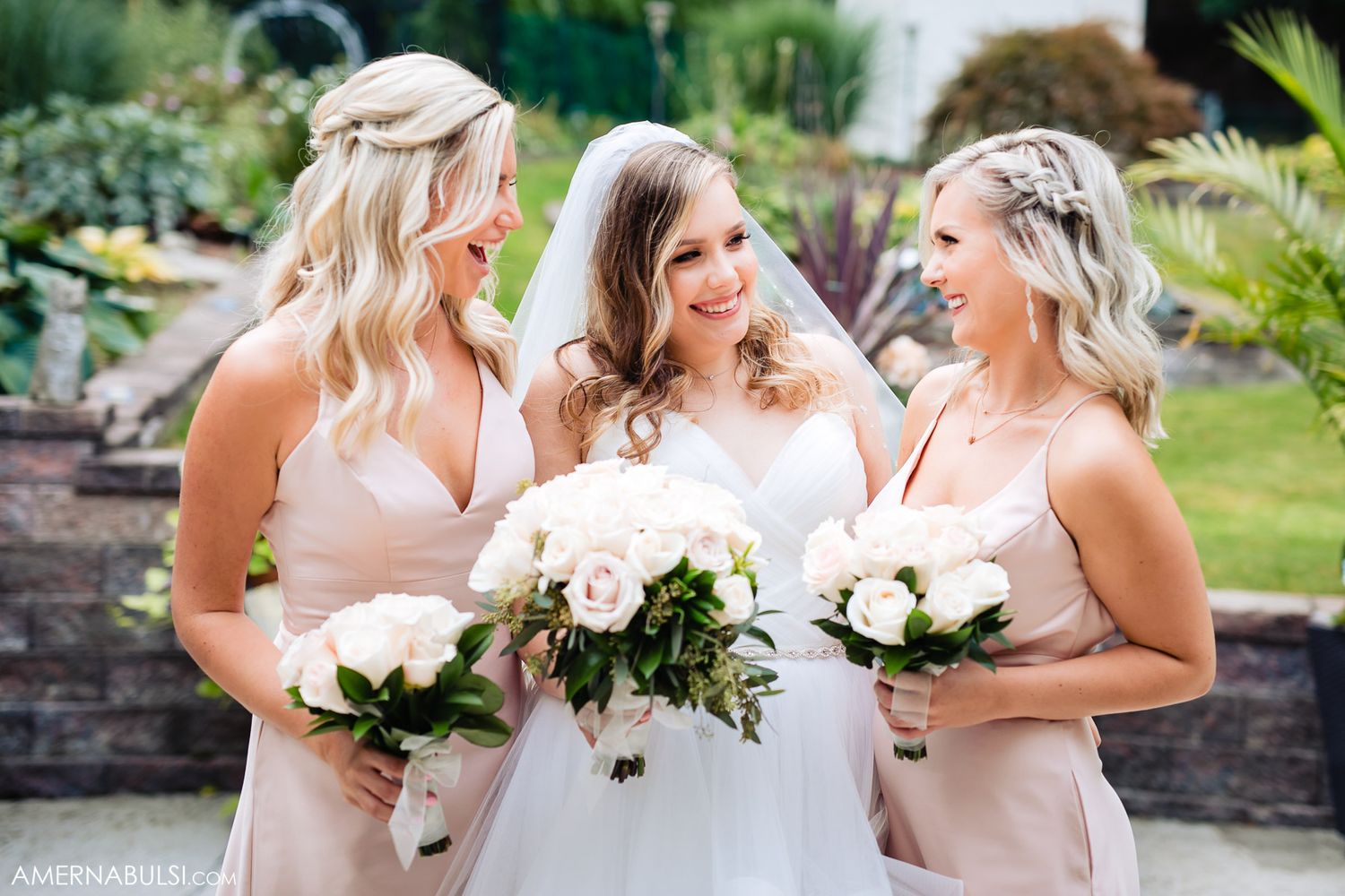 Bridesmaids in blush pink dresses share joyful moments while holding white floral bouquets in an outdoor garden setting.