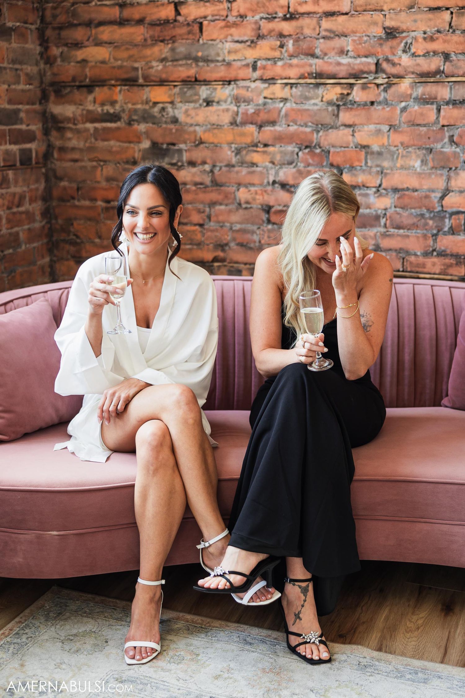 Two women in formal attire share drinks while seated on pink velvet couch.
