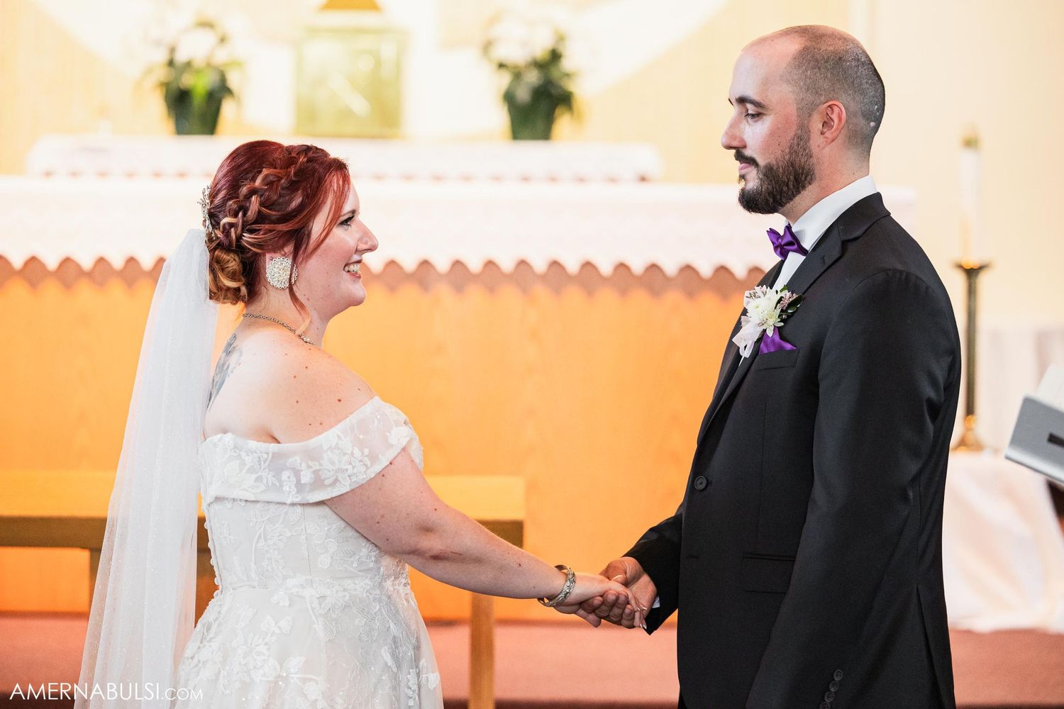 Wedding couple holding hands and smiling during marriage ceremony in church with orange walls.