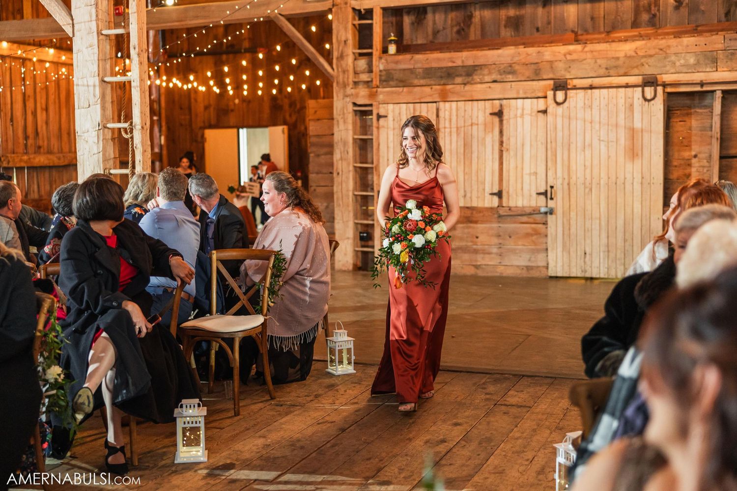 Wedding attendant in deep red attire walks through rustic barn venue with string lights and wooden beams.