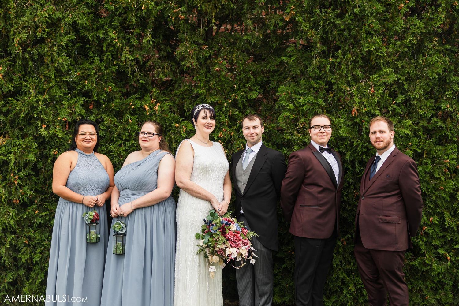 Wedding party in mix of burgundy suits and blue dresses poses against lush greenery.