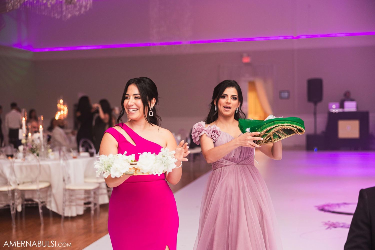 Two bridesmaids in pink dresses hold drinks during a wedding reception with purple lighting.