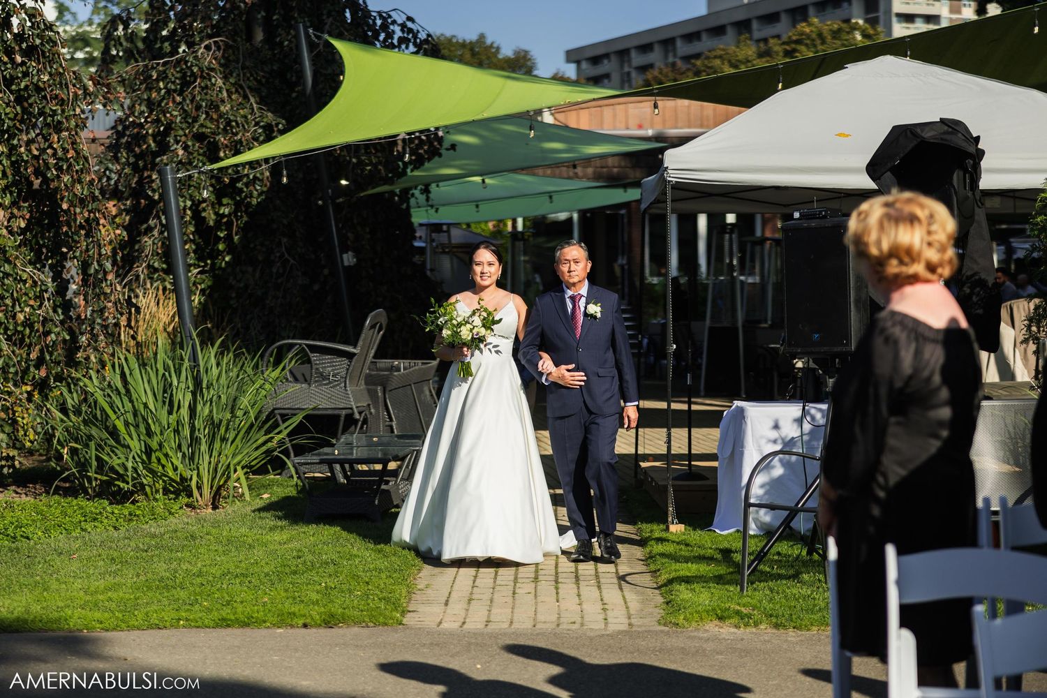 Outdoor wedding ceremony under a green shade sail with white chairs on a garden pathway.