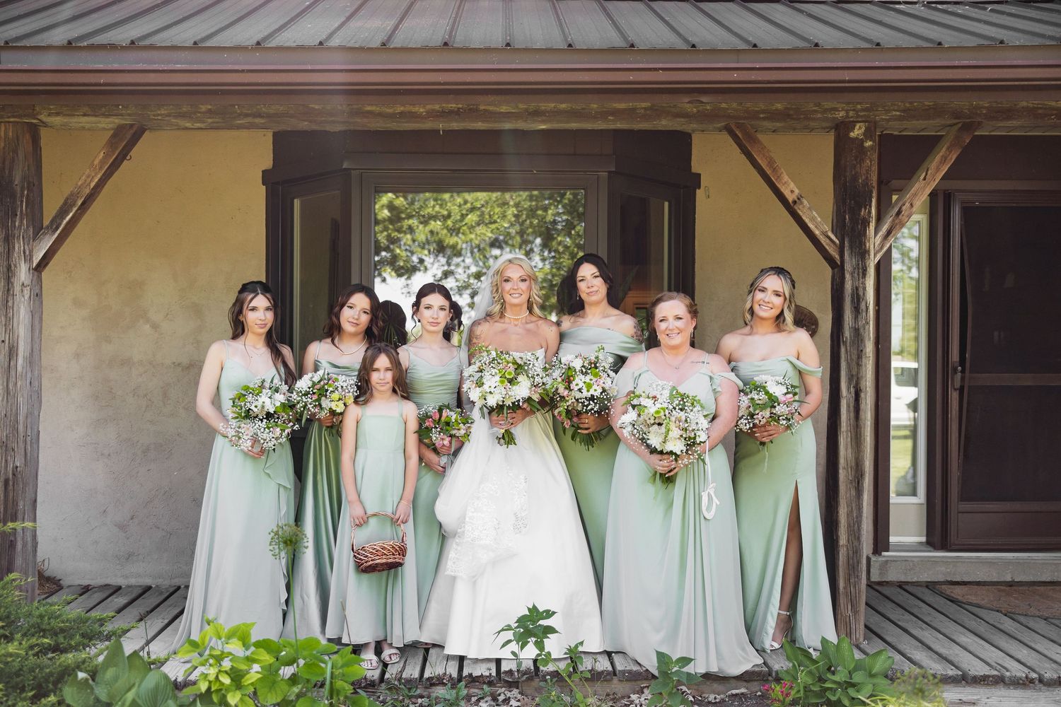 Bride in white gown poses with bridesmaids in sage green dresses holding floral bouquets outdoors.