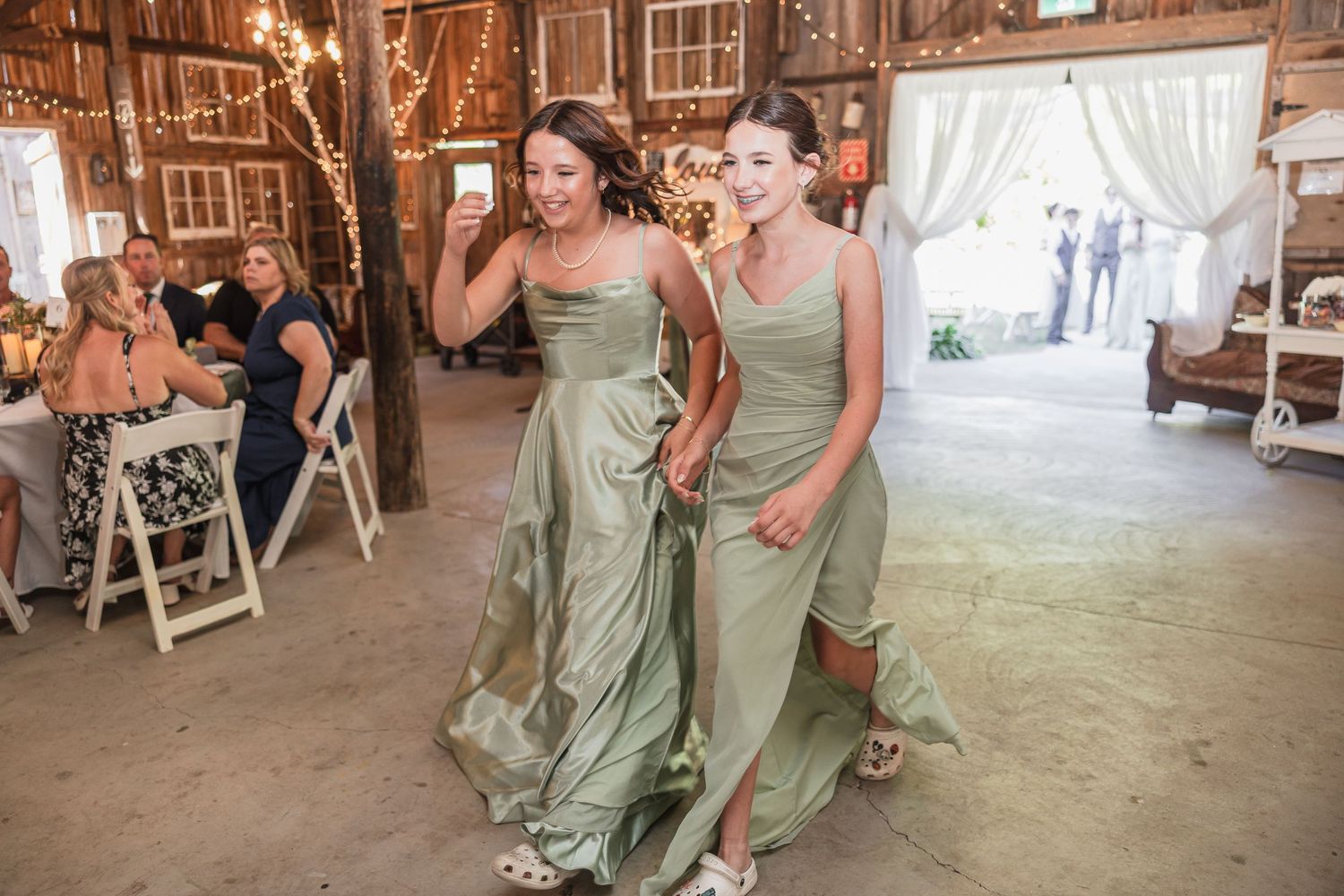 Two bridesmaids in sage green gowns walk through a rustic barn wedding venue with string lights.
