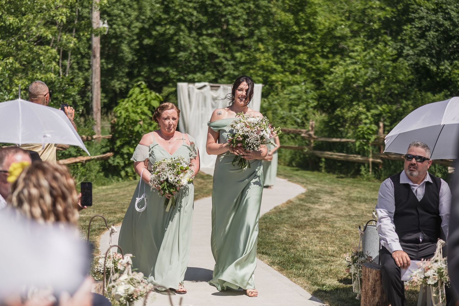Two bridesmaids in sage green gowns walk down an outdoor wedding aisle holding white bouquets.