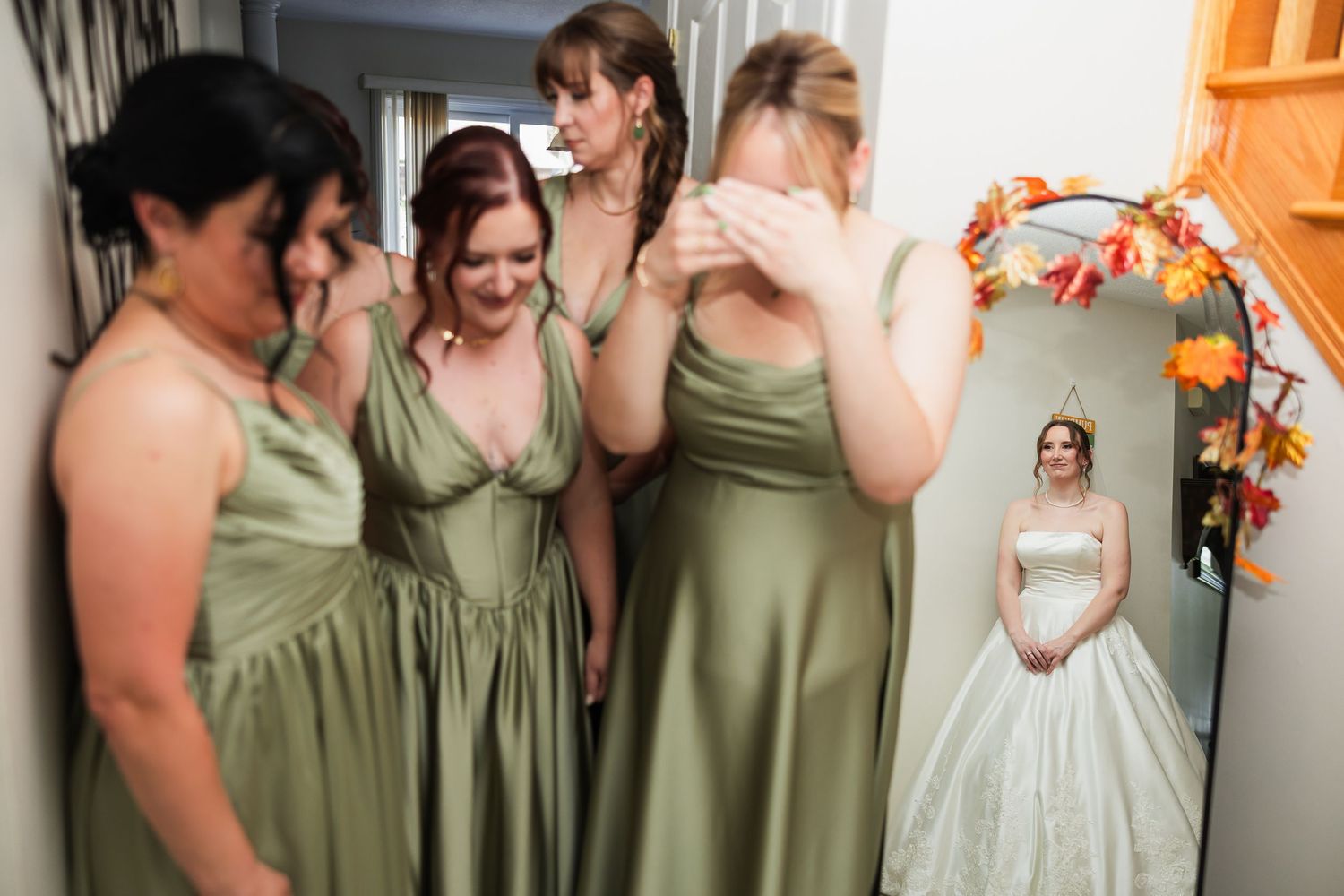 Bridesmaids in sage green dresses react emotionally as bride in white ballgown is reflected in mirror.