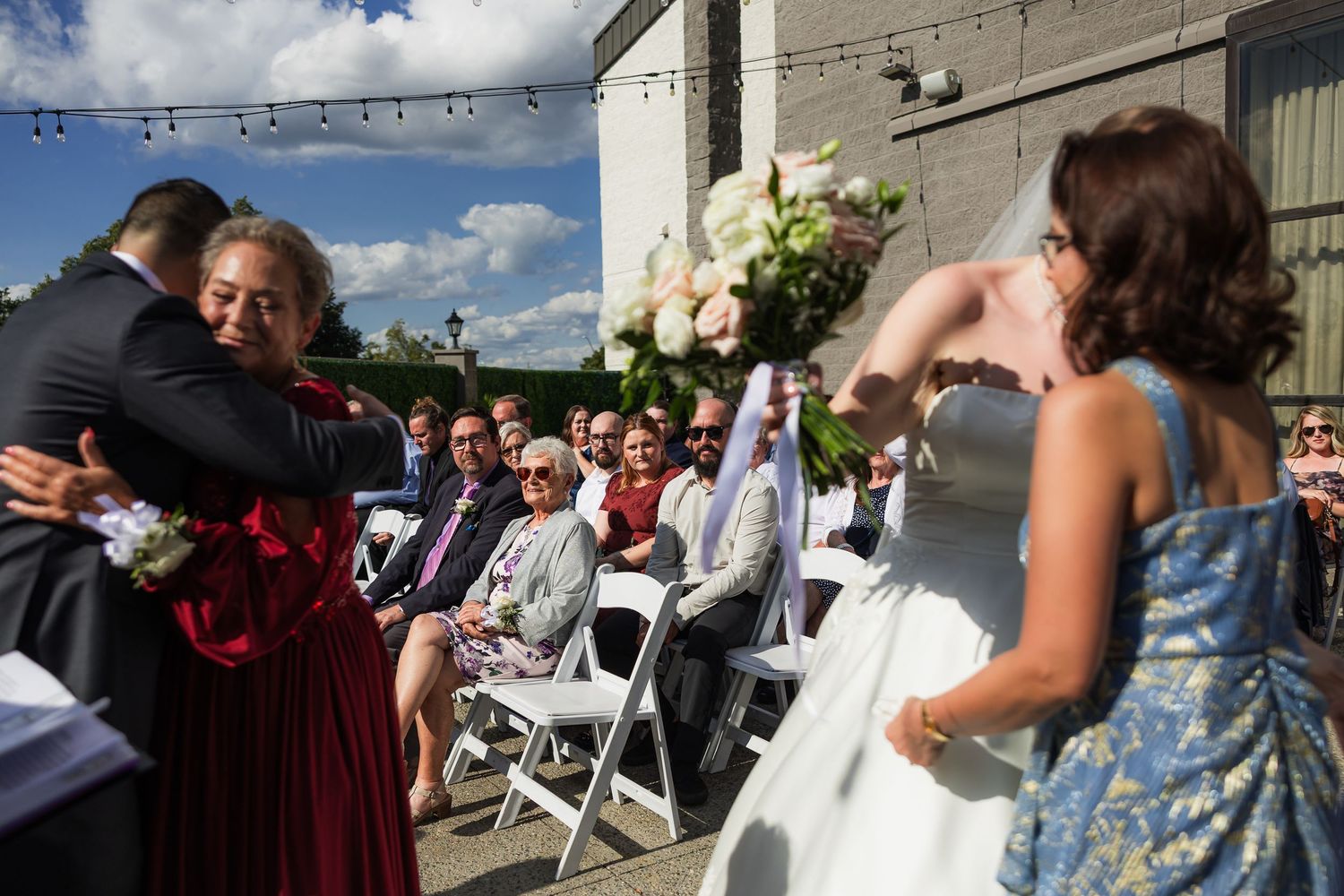 Outdoor wedding ceremony with guests seated as bride walks down aisle and groom embraces a woman in red dress.