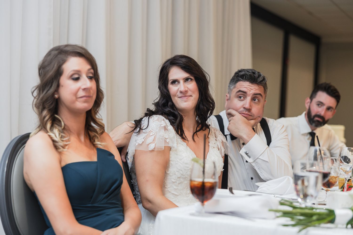 Wedding guests react with amusement at the reception table, including bride in lace gown and man in white jacket.