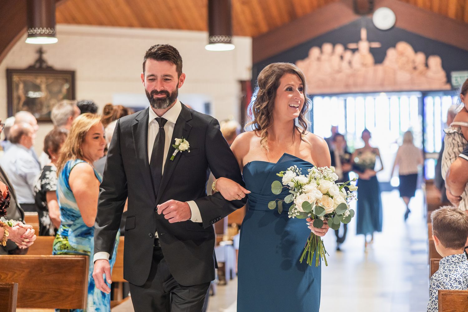 Groomsman escorts bridesmaid in blue dress down church aisle during wedding ceremony.