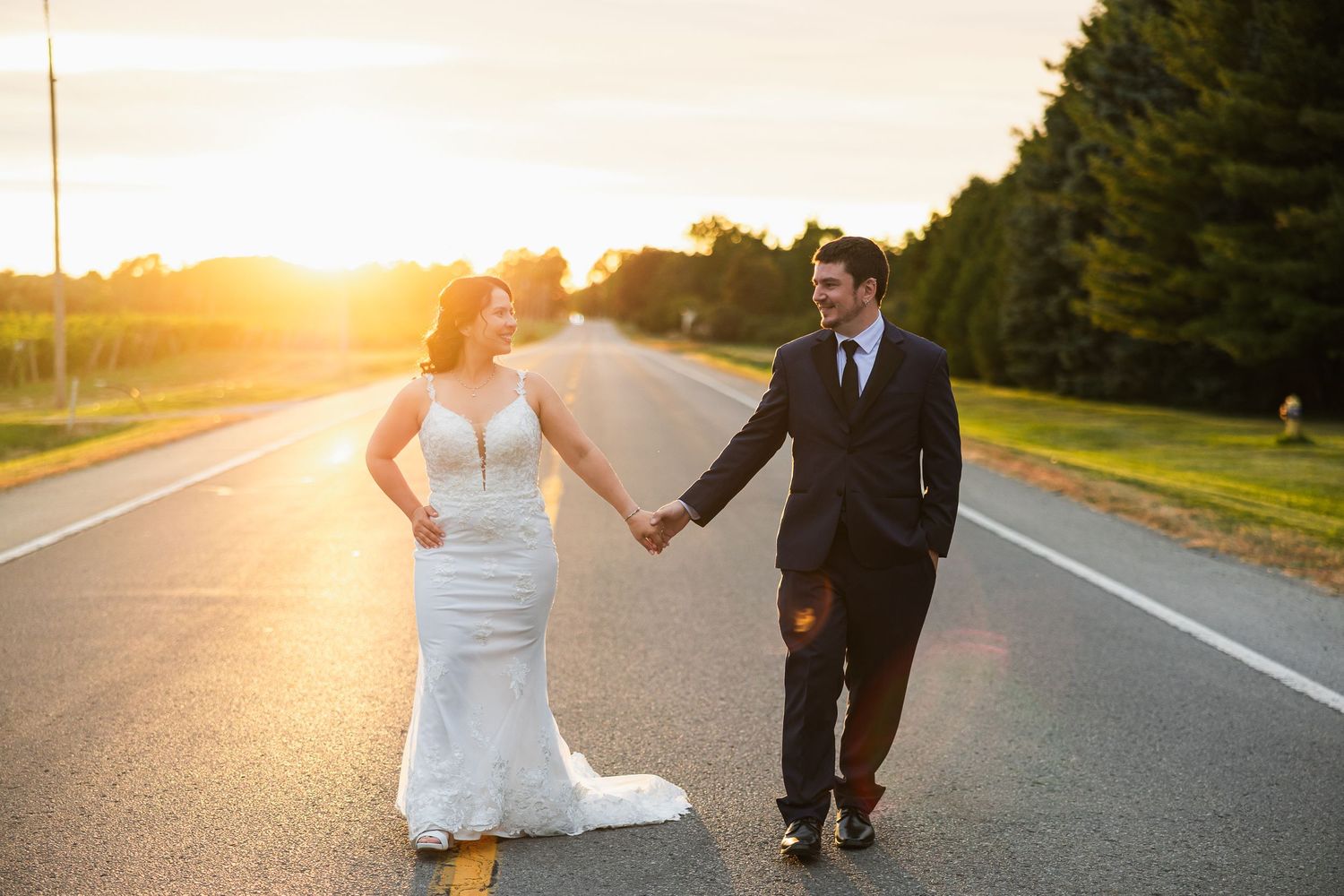 Bride and groom hold hands on an open road at golden sunset during romantic outdoor wedding portrait session.
