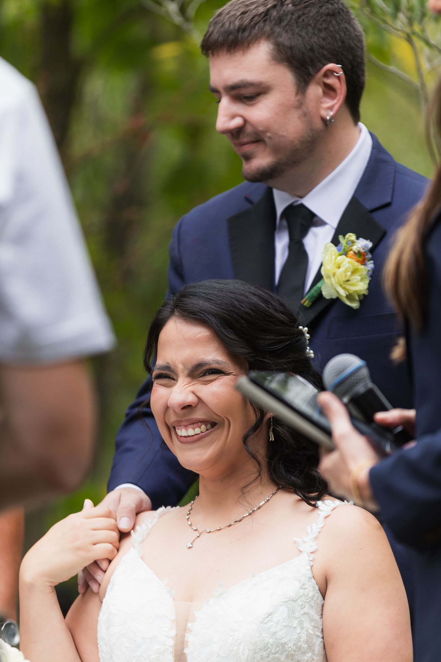 Bride smiles joyfully during outdoor wedding ceremony as groom stands behind her in a navy suit.