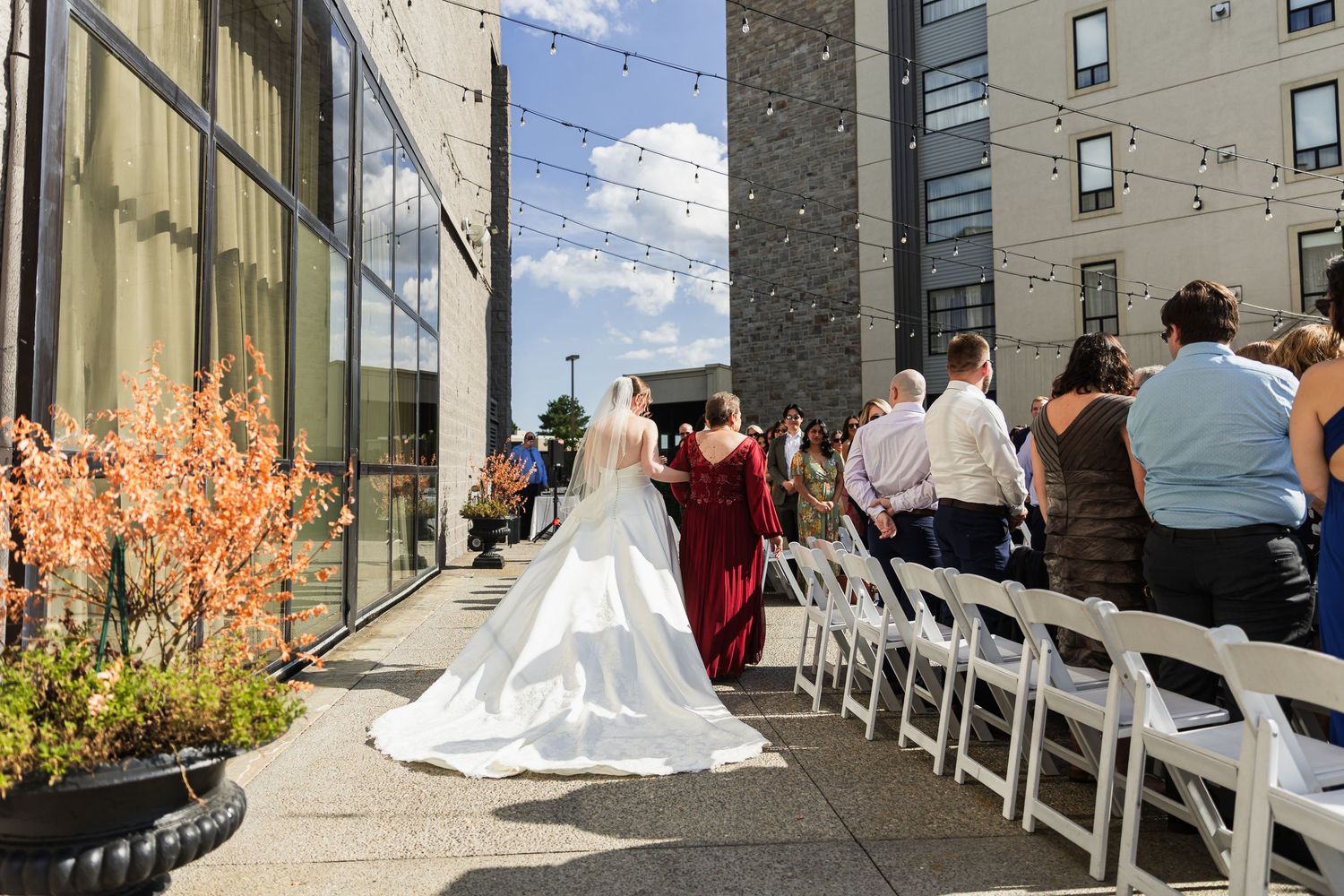 Bride in white gown and veil walks down outdoor aisle with guests seated in white chairs.