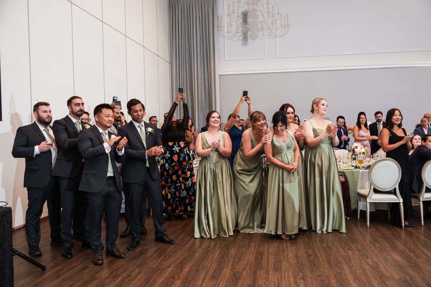 Wedding party in elegant venue, groomsmen in suits and bridesmaids in sage green gowns clapping and celebrating.