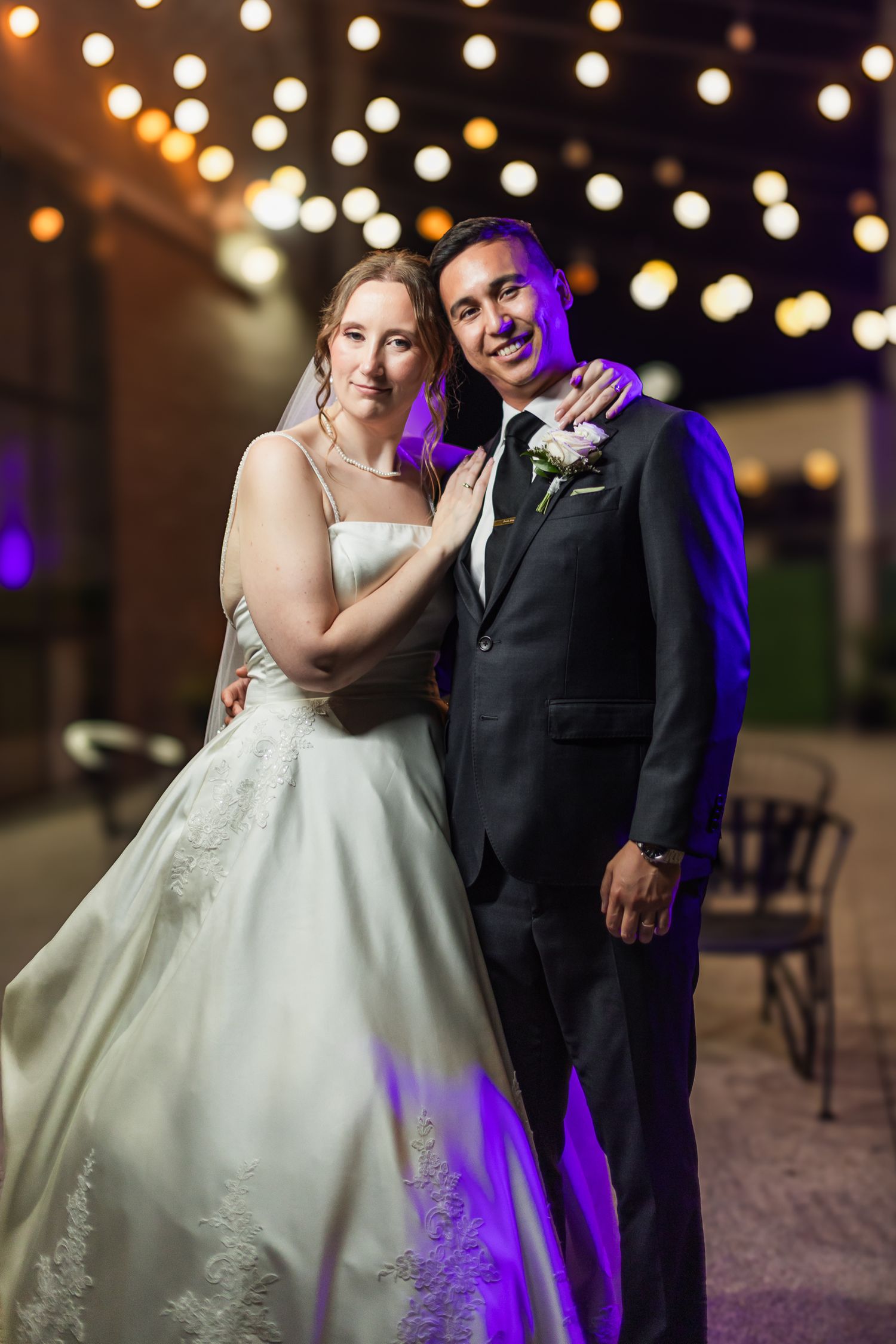 Bride in white lace gown and groom in black suit pose under string lights at night wedding reception.