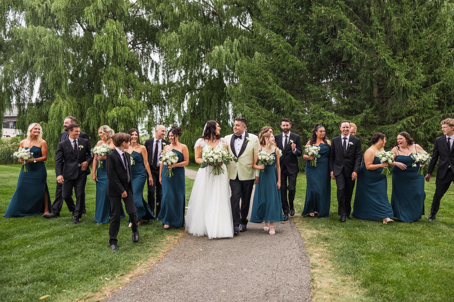 Wedding party walks together outdoors, bridesmaids in teal gowns and groomsmen in black suits with bride and groom.
