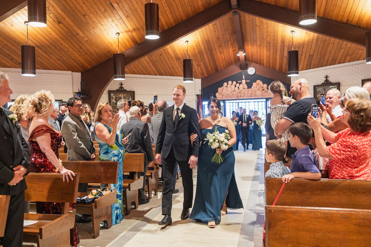 Groomsman walks a bridesmaid in a blue gown down the aisle of a warmly lit Catholic church wedding.