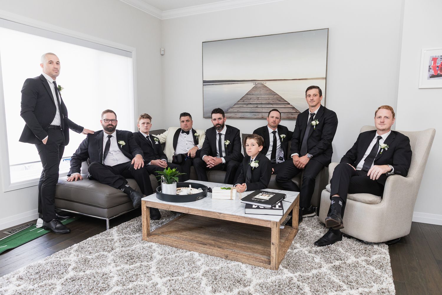 Groom and groomsmen in black tuxedos posing together in a modern living room before a wedding ceremony.