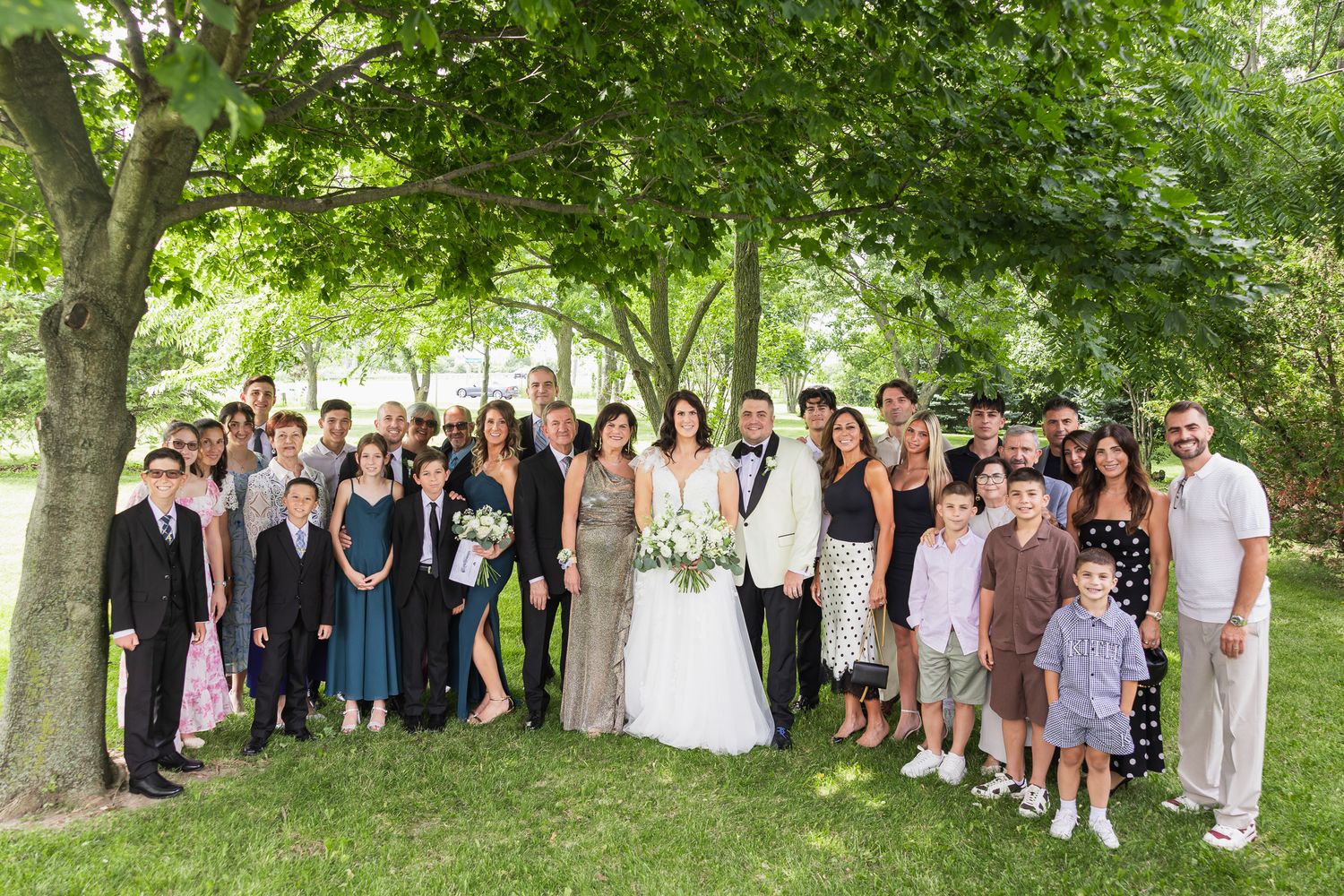 Large wedding family group photo with bride and groom surrounded by guests under trees.